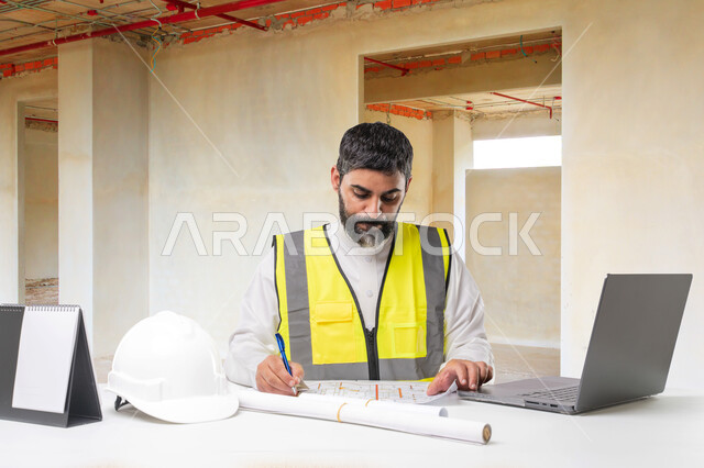 The concept of project management and auditing, working in the engineering sector, the role of architecture in the development of Saudi society, a close-up portrait of a Saudi Arabian Gulf engineer auditing and reviewing construction plans, sitting at his desk in front of an architectural background and using a laptop computer