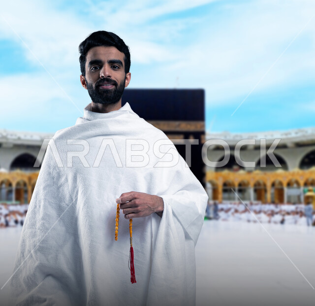 أداء مناسك الحج و العمرة, أماكن و معالم دينة إسلامية مقدسة في المملكة العربية السعودية, العبادة و التقرب إلى الله,صورة مقربة لرجل عربي خليجي سعودي يرتدي ملابس الاحرام يقف مبتسما امام الكعبه الشريفه ممسكا بمسبحته ذات الحبات الحمراء