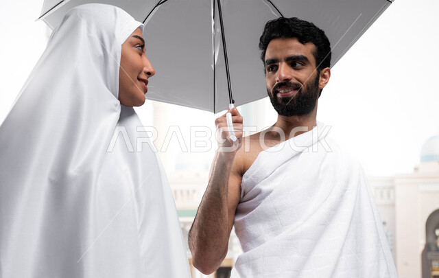 أداء مناسك الحج و العمرة, أماكن و معالم دينة إسلامية مقدسة في المملكة العربية السعودية, العبادة و التقرب إلى الله, صورة مقربة لزوجان عربيان خليجيان سعوديان مبتسمان يرتديان ملابس الاحرام يمسكان بمظلتهما الشمسيه استعدادا لاداء مناسك الحج
