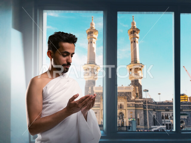The concept of the pillars of Islam, performing the rituals of Hajj and Umrah, supplication and prayer to the Lord of the servants, places and landmarks of the sacred Islamic religion in the Kingdom of Saudi Arabia, a close-up image of a Saudi Arabian Gulf man wearing the Ihram clothes, supplicating in front of the Holy House of God in Mecca
