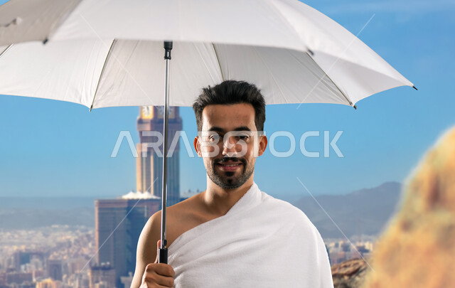 العبادة و التقرب إلى الله, أداء مناسك الحج و العمرة, أماكن و معالم دينة إسلامية مقدسة في المملكة العربية السعودية, صورة مقربة  لرجل عربي خليجي سعودي يرتدي ملابس الاحرام يقف مبتسما امام برج الساعه بمكه المكرمه ممسكا بمظلته البيضاء