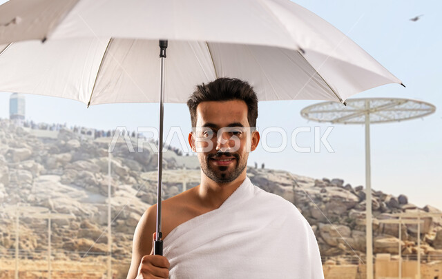 Sacred Islamic places and landmarks in the Kingdom of Saudi Arabia, performing Hajj and Umrah rituals, worship and drawing closer to God, a close-up of a Saudi Arabian Gulf man wearing Ihram clothes standing smiling in front of Mount Arafat holding his white umbrella