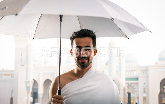 أداء مناسك الحج و العمرة, أماكن و معالم دينة إسلامية مقدسة في المملكة العربية السعودية, العبادة و التقرب إلى الله,صورة مقربة لرجل عربي خليجي سعودي يرتدي ملابس الاحرام يقف مبتسما امام المسجد الحرام ممسكا بمظلته البيضاء