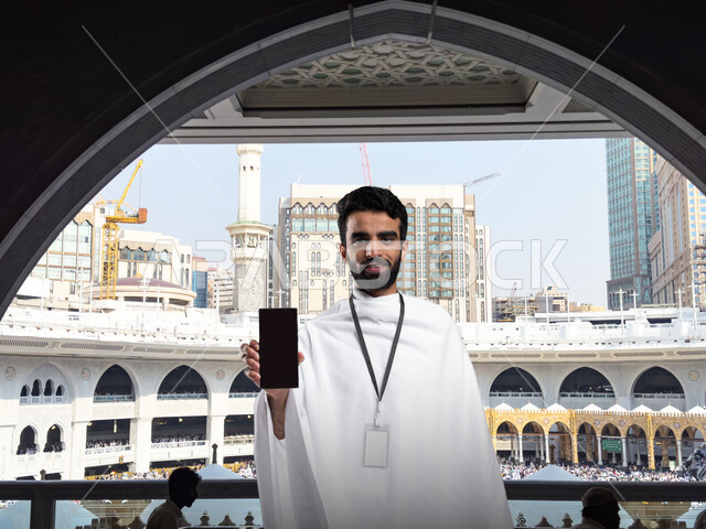 أداء مناسك الحج و العمرة, أماكن و معالم دينة إسلامية مقدسة في المملكة العربية السعودية, العبادة و التقرب إلى الله, صورة مقربة لرجل عربي خليجي سعودي يرتدي ملابس الاحرام يمسك بيده جهاز الهاتف المحمول بشاشه سوداء فارغه من امام الكعبه المشرفه بمكه المكرمه