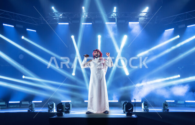 A Saudi Arabian Gulf man standing on the stage singing and encouraging, gestures of the face and hands indicating enthusiasm, highlighting the stage, a concert, and entertainment.