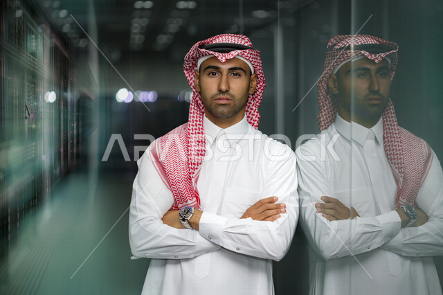 رجل عربي خليجي سعودي يقف مكتف اليدين داخل مقر الشركة ، موظف سعودي خليجي يرتدي الزي السعودي التقليدي ، شركة سعودية، بيئة عمل ، أعمال تجارية