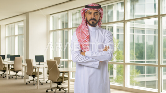 The concept of elegance, masculinity and interest in external appearance, a smiling Saudi Arabian Gulf man wearing a traditional shemagh and thobe, standing with his hands crossed in self-confidence gestures, a large glass facade background, working in companies in the Kingdom of Saudi Arabia
