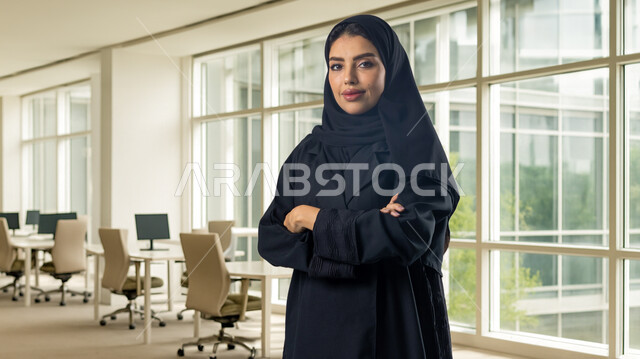 Saudi women's office jobs and professions, managing and completing tasks using a modern and advanced technical device, a smiling Saudi Arabian Gulf woman wearing a hijab and abaya standing with crossed hands in gestures of self-confidence at the company headquarters, expressions of pleasure and happiness