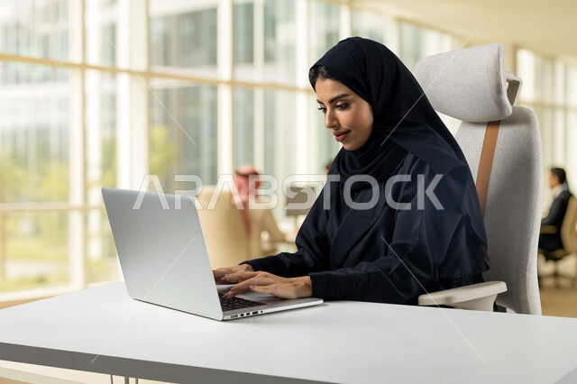 مهنة ووظيفة مكتبية، ادارة وانجاز المهام باستخدام جهاز تقني حديث ومتطور, إمرأة عربية خليجية سعودية مبتسمة ترتدي الحجاب والعباءة تعمل على الحاسوب المحمول في مقر الشركة, ايماءات وتعابير السرور والسعادة