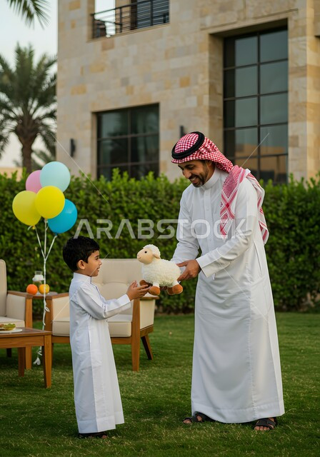 التسامر والضحك مع الابناء في عيد الاضحى المبارك، رجل عربي خليجي سعودي يرتدي الشماغ والثوب التقليدي يعطي ابنه دمية على شكل خاروف عيد الاضحى، التزيين في الاعياد والمناسبات العائليه, ايماءات السعادة والسرور