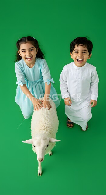 قربان واضحية عيد الأضحى المبارك، الاحتفال بالمناسبات والأعياد الدينية الإسلامية، بورتريه لولد عربي خليجي سعودي مبتسم يرتدي الثوب التقليدي يقف مع اخته بجانب خاروف أبيض لطيف،  خلفية خضراء