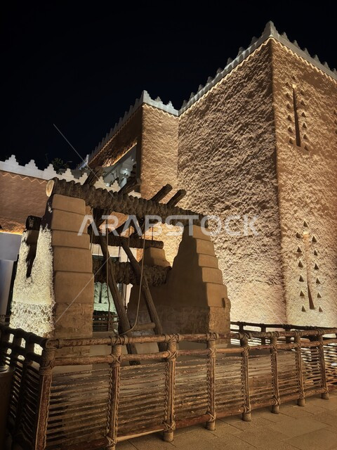 بئر تقليدي خشبي أمام مبنى طيني تراثي مضاء ليلا في البجيري في حي الطريف، المباني والبيوت الطينية في الدرعية القديمة بمدينة الرياض، المواقع الأثرية والسياحية في المملكة، التصاميم و الفنون الهندسية المعمارية للمباني الطينية على الطراز النجدي