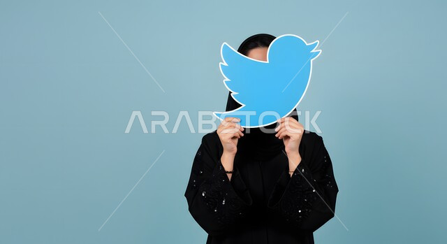 Mobile apps, social media browsing, close-up portrait of a Saudi Arabian Gulf woman wearing hijab and abaya holding Twitter app logo, social media icon, blue background