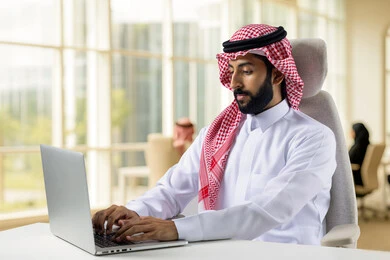  مفهوم دمج العمل مع التقنيه والتكنولوجيا, إنجاز مهام العمل عبر جهاز الابتوب, رجل عربي خليجي سعودي مبتسم يرتدي الثوب والشماغ التقليدي يعمل على الحاسب المحمول  بإيماءات السرور في مقر الشركة