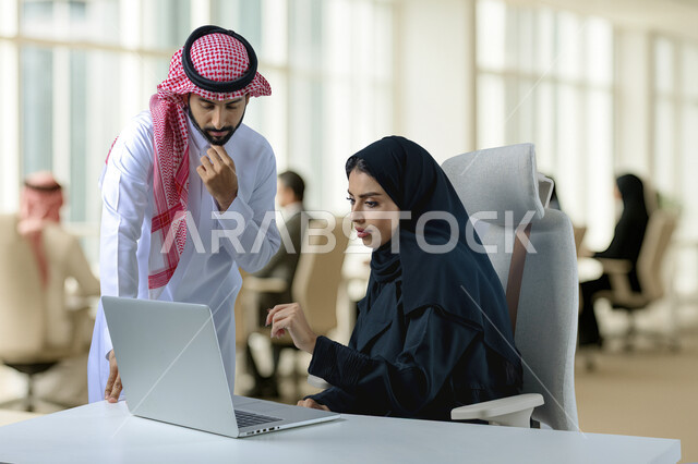 بيئة العمل المريحة في الشركات السعودية، الحوار و النقاش بين الزملاء، تبادل الخبرات والمعلومات لتحقيق الاهداف, موظفان عربيان خليجيان سعوديان يجلسان على مكتبهم داخل مقر الشركة، استخدام الحاسب والتقنية في مجال الاعمال