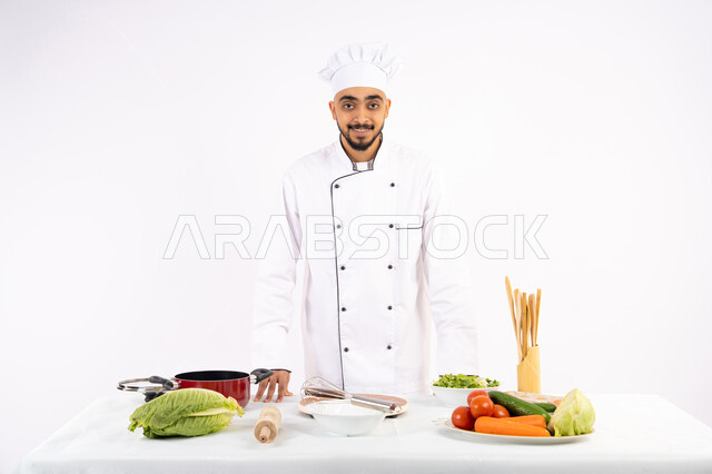 Looking at the camera with confident gestures, preparing to prepare salads and healthy dishes, concept of following a healthy diet, portrait of an Emirati Gulf Arab chef wearing his professional uniform standing in front of a table with a variety of fresh vegetables, white background