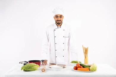 النظر إلى الكاميرا بإيماءات الثقة بالنفس، الاستعداد لإعداد السلطات والأطباق الصحية، مفهوم اتباع النظام الغذائي الصحي، بورتريه لشيف عربي خليجي اماراتي يرتدي الزي الخاص بمزاولة المهنة يقف أمام طاولة عليها مجموعة من الخضراوات الطازجة، خلفية بيضاء