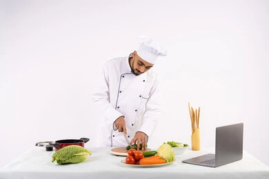 التدرب على مهارة الطهي وإعداد الطعام عبر الحاسب المحمول، بورتريه لرجل عربي خليجي اماراتي يرتدي زي العمل يقوم بتحضير وصفة طبق صحي مفيد من الانترنت، شيف سعودي يقوم بتقطيع الخضراوات الطازجة، استخدام المكونات والمقادير المناسبة للوجبة، خلفية بيضاء