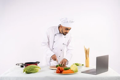 استخدام المكونات والمقادير المناسبة للوجبة، التدرب على مهارة الطهي وإعداد الطعام عبر الحاسب المحمول، بورتريه لرجل عربي خليجي اماراتي يرتدي زي العمل يقوم بتحضير وصفة طبق صحي مفيد من الانترنت، شيف سعودي يقوم بتقطيع الخضراوات الطازجة، خلفية بيضاء