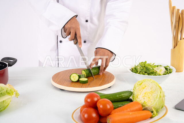 شغف التدرب على مهارة الطهي وإعداد الطعام، احترافية تقطيع الخضار، بورتريه مقرب لأيدي رجل عربي خليجي اماراتي يرتدي زي العمل يحضر طبق صحي مفيد، شيف سعودي يقوم بتحضير سلطة الخضراوات الطازجة، استخدام المكونات والمقادير المناسبة للوجبة، خلفية بيضاء