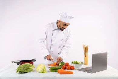 استخدام المكونات والمقادير المناسبة للوجبة، التدرب على مهارة الطهي وإعداد الطعام عبر الحاسب المحمول، بورتريه لرجل عربي خليجي اماراتي يرتدي زي العمل يقوم بتحضير وصفة طبق صحي مفيد من الانترنت، شيف سعودي يقوم بتقطيع الخضراوات الطازجة، خلفية بيضاء
