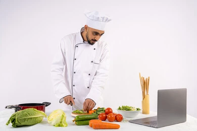 تعلم فنون الطبخ، التدرب على إعداد الطعام عبر الحاسب المحمول، بورتريه لرجل عربي خليجي اماراتي يرتدي زي العمل يقوم بتحضير وصفة طبق صحي مفيد من الانترنت، شيف سعودي يقوم بتقطيع الخضراوات الطازجة، استخدام المكونات والمقادير المناسبة للوجبة، خلفية بيضاء