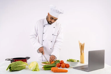 الخضراوات والمقادير المناسبة للوجبة، التدرب على مهارة الطهي وإعداد الطعام عبر الحاسب المحمول، بورتريه لرجل عربي خليجي اماراتي يرتدي زي العمل يقوم بتحضير وصفة طبق صحي مفيد من الانترنت، شيف سعودي يقوم بتقطيع الخضراوات الطازجة، خلفية بيضاء