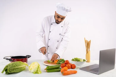 اتباع نظام غذائي صحي، التدرب على مهارة الطهي عبر الحاسب المحمول، بورتريه لرجل عربي خليجي اماراتي يرتدي زي العمل يقوم بتحضير وصفة طبق صحي مفيد من الانترنت، شيف سعودي يقوم بتقطيع الخضراوات الطازجة، استخدام المكونات والمقادير المناسبة للوجبة، خلفية بيضاء