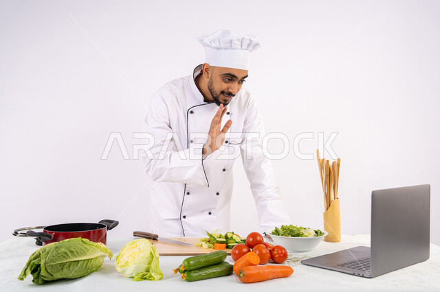 Greeting and greeting, learning and practicing the art of cooking through websites using a computer, raising community awareness about preparing healthy food, portrait of an Emirati Gulf Arab man wearing work clothes filming and displaying cooking via laptop, Saudi chef preparing a healthy recipe via live broadcast, white background