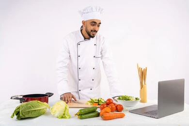 مزاولة مهنة الطباخ، التدرب على مهارة الطهي عبر الحاسب المحمول، بورتريه لرجل عربي خليجي اماراتي يرتدي زي العمل يقوم بتحضير وصفة طبق صحي مفيد من الانترنت، شيف سعودي يقوم بتقطيع الخضراوات الطازجة، استخدام المكونات والمقادير المناسبة للوجبة، خلفية بيضاء
