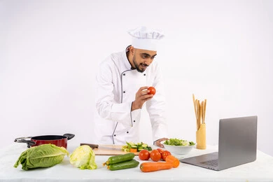 Applying and learning the art of cooking through websites using a computer, raising community awareness of preparing healthy food, a portrait of an Arab Gulf Emirati man wearing work clothes filming and displaying cooking via laptop, a Saudi chef explaining the ingredients and quantities required via a live broadcast, white background