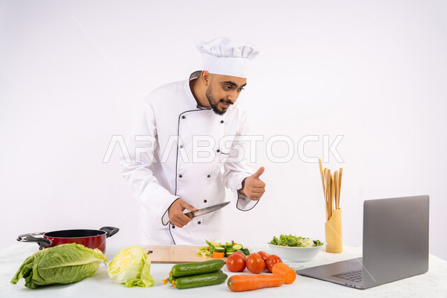 Thumbs up gesture, learning cooking arts through websites via computer, educating society about preparing healthy food, portrait of an Emirati Gulf Arab man wearing work clothes filming and showing cooking via laptop, Saudi chef preparing a recipe via live broadcast, white background