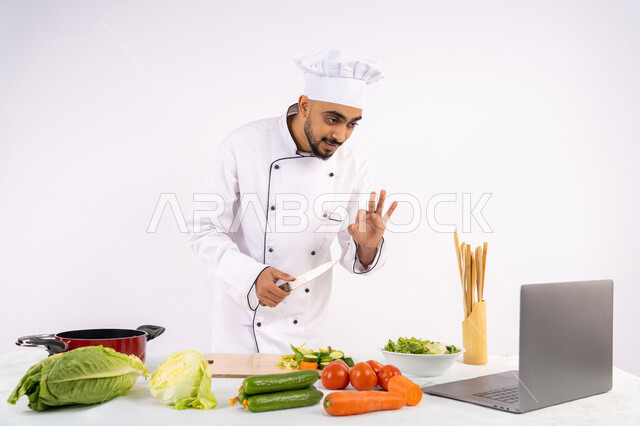 Hand gestures indicating admiration, learning and applying culinary arts through websites using a computer, raising community awareness about preparing healthy food, portrait of an Emirati Gulf Arab man wearing work clothes filming and displaying cooking via laptop, Saudi chef preparing a healthy recipe via live broadcast, white background