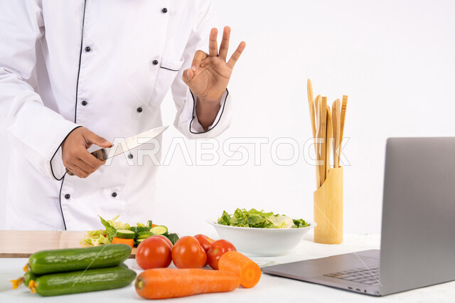 Pointing with thumbs up gestures, learning and applying culinary arts through websites using a computer, raising community awareness about preparing healthy food, portrait of an Emirati Gulf Arab man wearing work clothes filming and displaying cooking via laptop, Saudi chef preparing a healthy recipe via live broadcast, white background