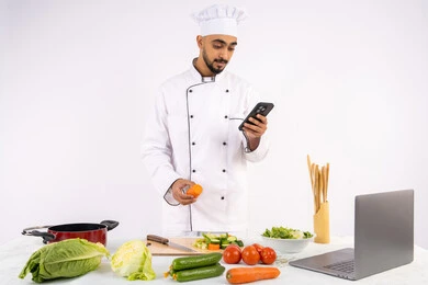 Community awareness of healthy food preparation, learning and applying culinary arts through websites via computer, portrait of an Emirati Gulf Arab man wearing work clothes using a mobile phone, Saudi chef preparing a recipe with healthy vegetables, white background