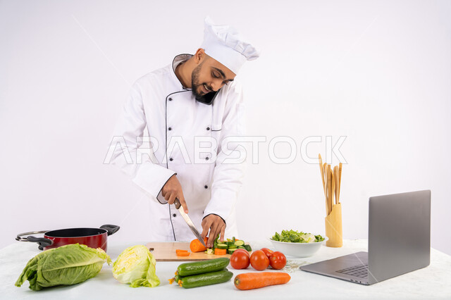 إجراء مكالمة هاتفية، التدرب على مهارة الطهي وإعداد الطعام عبر الحاسوب، بورتريه لرجل عربي خليجي اماراتي يرتدي زي العمل يقوم بتحضير وصفة طبق صحي مفيد من الانترنت، شيف سعودي يقوم بتقطيع الخضراوات الطازجة، استخدام المكونات والمقادير المناسبة للوجبة، خلفية بيضاء