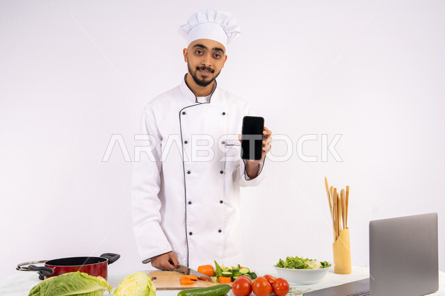 نشر الوعي في المجتمع بإعداد الطعام الصحي، تعلم وتطبيق فنون الطهي من خلال المواقع الإلكترونية باستخدام الحاسوب، بورتريه لرجل عربي خليجي اماراتي يرتدي زي العمل يقوم بعرض شاشة سوداء فارغة عبر الهاتف المحمول، شيف سعودي يحضر وصفة بالخضراوات الصحية، خلفية بيضاء