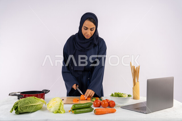 تعلم مهارة الطهي وإ عداد الطعام عبر الحاسب المحمول، بورتريه لسيدة عربية خليجية اماراتية محجبة  ترتدي عباءة تقوم بتحضير وصفة طبق صحي مفيد من الانترنت، ربة منزل سعودية تقوم بتقطيع الخضراوات الطازجة، استخدام المكونات والمقادير المناسبة للوجبة، خلفية بيضاء