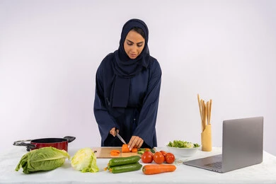 تعلم مهارة الطهي وإ عداد الطعام عبر الحاسب المحمول، بورتريه لسيدة عربية خليجية اماراتية محجبة  ترتدي عباءة تقوم بتحضير وصفة طبق صحي مفيد من الانترنت، ربة منزل سعودية تقوم بتقطيع الخضراوات الطازجة، استخدام المكونات والمقادير المناسبة للوجبة، خلفية بيضاء