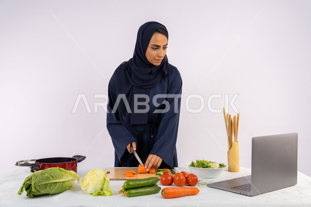 إعداد السلطات والاطباق الصحية، تعلم مهارة الطهي عبر الحاسب المحمول، بورتريه لسيدة عربية خليجية اماراتية محجبة  ترتدي عباءة تقوم بتحضير وصفة طبق صحي مفيد من الانترنت، ربة منزل سعودية تقوم بتقطيع الخضراوات الطازجة، خلفية بيضاء