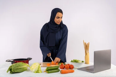 Preparing salads and healthy dishes, learning cooking skills via laptop, portrait of a veiled Emirati Gulf Arab woman wearing an abaya preparing a healthy dish recipe from the internet, a Saudi housewife cutting fresh vegetables, white background