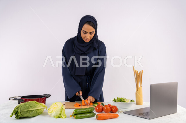 إتقان فنون الطبخ، تعلم إعداد الطعام عبر الحاسب المحمول، بورتريه لسيدة عربية خليجية اماراتية محجبة  ترتدي عباءة تقوم بتحضير وصفة طبق صحي مفيد من الانترنت، ربة منزل سعودية تقوم بتقطيع الخضراوات الطازجة، استخدام المكونات والمقادير المناسبة للوجبة، خلفية بيضاء