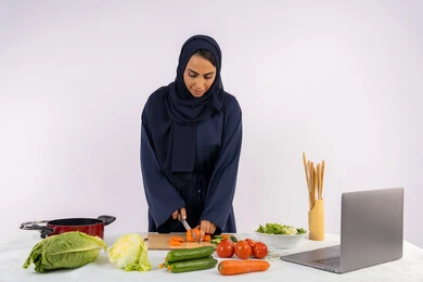 Mastering the art of cooking, learning to prepare food via laptop, portrait of a veiled Emirati Gulf Arab woman wearing an abaya preparing a healthy and useful dish recipe from the internet, a Saudi housewife chopping fresh vegetables, using the appropriate ingredients and quantities for the meal, white background