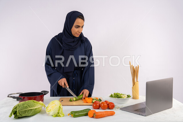 Publishing healthy recipes, preparing salads and healthy dishes, learning cooking skills via laptop, portrait of a veiled Emirati Gulf Arab woman wearing an abaya preparing a healthy and useful dish recipe from the internet, a Saudi housewife cutting fresh vegetables, white background