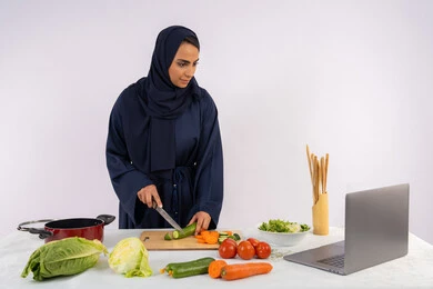 نشر الوصفات الصحية، إعداد السلطات والاطباق الصحية، تعلم مهارة الطهي عبر الحاسب المحمول، بورتريه لسيدة عربية خليجية اماراتية محجبة  ترتدي عباءة تقوم بتحضير وصفة طبق صحي مفيد من الانترنت، ربة منزل سعودية تقوم بتقطيع الخضراوات الطازجة، خلفية بيضاء