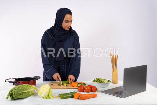 تعلم فنون الطبخ في الدورات التدريبيه عبر الحاسب المحمول، بورتريه لسيدة عربية خليجية اماراتية محجبة  ترتدي عباءة تقوم بتحضير وصفة طبق صحي مفيد من الانترنت، ربة منزل سعودية تقوم بتقطيع الخضراوات الطازجة، استخدام المكونات والمقادير المناسبة للوجبة، خلفية بيضاء