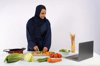 تعلم فنون الطبخ في الدورات التدريبيه عبر الحاسب المحمول، بورتريه لسيدة عربية خليجية اماراتية محجبة  ترتدي عباءة تقوم بتحضير وصفة طبق صحي مفيد من الانترنت، ربة منزل سعودية تقوم بتقطيع الخضراوات الطازجة، استخدام المكونات والمقادير المناسبة للوجبة، خلفية بيضاء