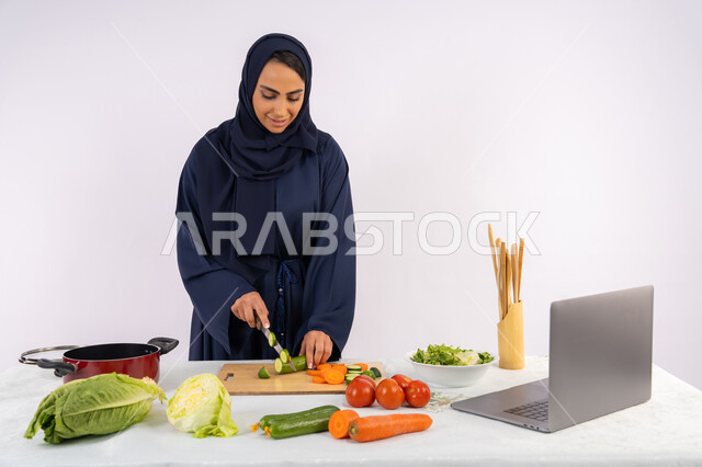 إعداد السلطات والاطباق الصحية، تعلم مهارة الطهي عبر الحاسب المحمول، بورتريه لسيدة عربية خليجية اماراتية محجبة  ترتدي عباءة تقوم بتحضير وصفة طبق صحي مفيد من الانترنت، ربة منزل سعودية تقوم بتقطيع الخضراوات الطازجة، خلفية بيضاء