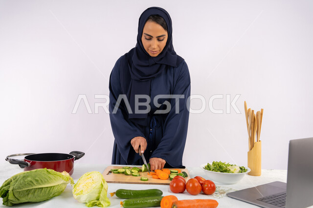Learning the skill of cooking and preparing food via laptop, portrait of a veiled Emirati Gulf Arab woman wearing an abaya preparing a recipe for a healthy and useful dish from the internet, a Saudi housewife cutting fresh vegetables, using the appropriate ingredients and quantities for the meal, white background