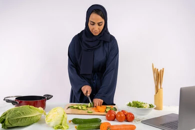 تعلم مهارة الطهي وإ عداد الطعام عبر الحاسب المحمول، بورتريه لسيدة عربية خليجية اماراتية محجبة  ترتدي عباءة تقوم بتحضير وصفة طبق صحي مفيد من الانترنت، ربة منزل سعودية تقوم بتقطيع الخضراوات الطازجة، استخدام المكونات والمقادير المناسبة للوجبة، خلفية بيضاء